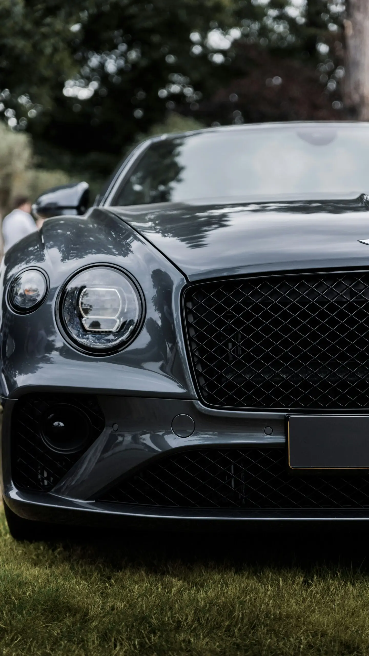 Elegant Bentley Luxury Car Front View Outdoors on Green Grass Close-up of a dark grey Bentley car's front grille and headlights, parked on green grass outdoors.