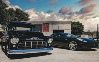 Chevy Truck Ferrari Ff Jfm A vintage black pickup truck parked next to a sleek black sports car on a sunny day with clouds.
