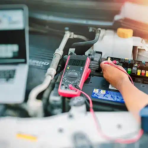 Car Battery Testing With Multimeter and Laptop Technician using a multimeter to test a car battery, with a laptop nearby.