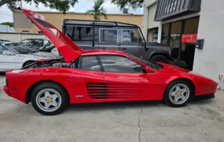 Red Ferrari Testarossa With Rear Hood Open Vibrant red Ferrari Testarossa sports car with its rear engine cover open, parked on concrete with other cars in background