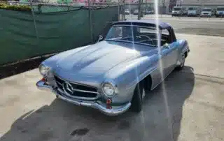 Vintage Silver Mercedes-Benz 190SL Convertible on Pavement A classic silver Mercedes-Benz 190SL convertible parked outdoors on a paved surface, with a green fence in the background.