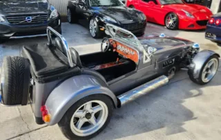 Custom Grey Three-Wheeled Car with Side Exhaust A custom-built grey three-wheeled car with a side exhaust pipe and a spare tire mounted on the rear.