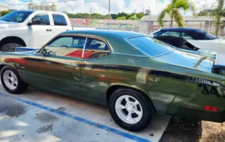Vintage Green Plymouth Duster Muscle Car Parked Outdoors Olive green Plymouth Duster muscle car with black side stripe and white custom wheels parked in a lot beside a white truck.