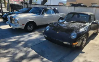 Classic Cars: Rolls-Royce And Porsche Parked Outdoors A white Rolls-Royce and a black Porsche 911 parked on a concrete surface, with trees and a building in the background.